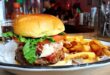 Close-up of a juicy classic burger topped with lettuce, tomato, and shaved parmesan, served alongside a generous portion of golden fries. The burger is perfectly placed on a white plate, captured in a bustling diner setting that enhances the casual yet inviting atmosphere of the eatery